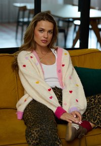 Biała dziergana kardigan z kolorowymi wzorami serduszek i różowymi wykończeniami, w połączeniu z spodniami w panterkę, na tle żółtej kanapy.