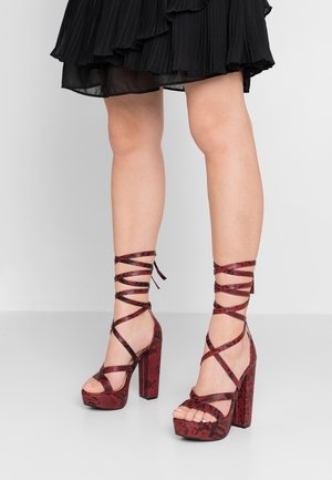 Gambe che indossano tacchi con plateau in pelle di serpente rossa con cinturini alla caviglia, abbinati a una gonna nera a pieghe su uno sfondo grigio.