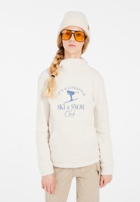 Felpa color crema con collo alto, tasca frontale e testo grafico blu. Abbinata a un berretto lavorato a maglia color crema e occhiali con lenti gialle.