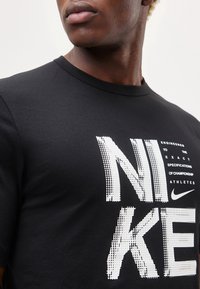 Κοντινό ενός ατόμου που φοράει μαύρο μπλουζάκι Nike με μεγάλο λευκό κείμενο Nike και μικρό σύνθημα στο στήθος.