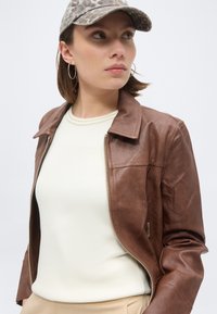 Γυναίκα με σκουλαρίκια κρίκους, καπέλο καμουφλάζ, λευκό τοπ και καφέ δερμάτινο μπουφάν, κοιτάζει προς τα πλάγια με φόντο ανοιχτόχρωμο.