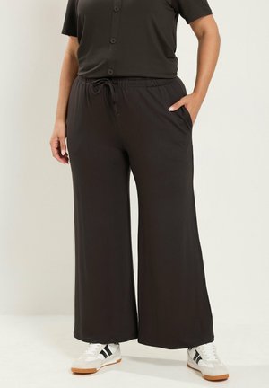 Persoon die losse zwarte broek draagt met een elastische tailleband en witte sneakers met zwarte strepen, staand met één hand in de zak.