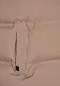 Primo piano di una tasca di una giacca trapuntata beige, con un piccolo logo quadrato nero vicino al bordo inferiore della pattella della tasca.