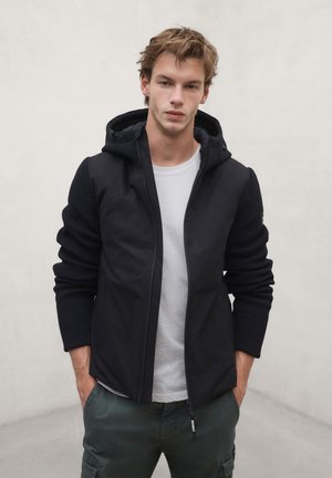 Young man with light brown hair wears a black hooded jacket over a white shirt and green cargo pants, standing with hands in pockets.