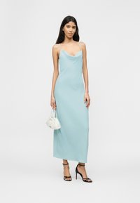 Γυναίκα με ανοιχτό γαλάζιο αμάνικο φόρεμα, κρατώντας μια μικρή λευκή τσάντα και φορώντας μαύρα πέδιλα με ψηλό τακούνι, σε λευκό φόντο.