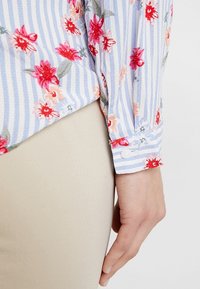 Ljusblå och vit randig skjorta med röd och rosa blommig broderi, knappade ärmslut och texturerad tyg. Bärs med beige byxor.