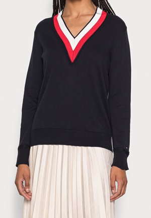 Pull noir à manches longues avec col en V et rayures rouges, blanches et bleu marine, associé à une jupe plissée blanche sur une personne aux cheveux tressés.