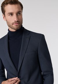 Marineblauer strukturierter Blazer mit Revers und einem Knopfverschluss, getragen über einem passenden marineblauen Rollkragenpullover. Dezentes Markendetail am Revers.