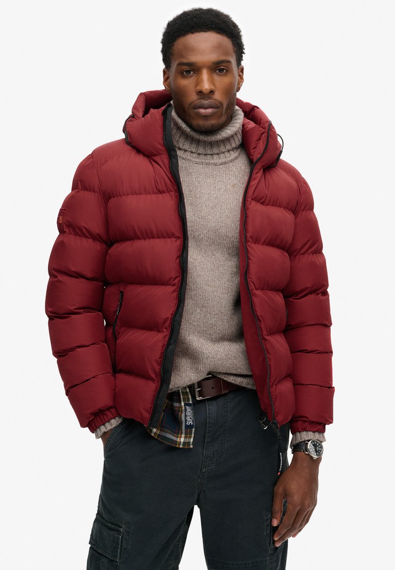 Doudoune rouge avec capuche, poignets côtelés et fermeture zippée, portée sur un pull col roulé beige et un pantalon cargo noir.