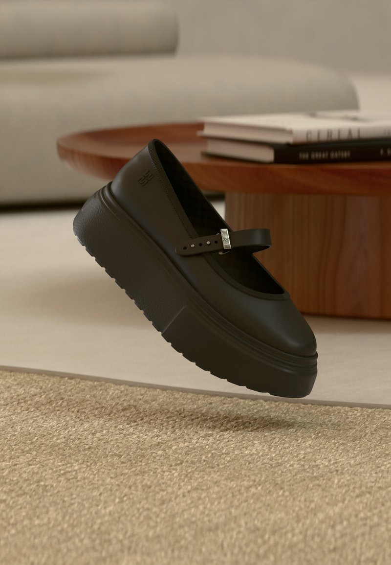 Chaussure Mary Jane à plateforme noire avec bride ajustable flottant au-dessus d'un tapis beige, table en bois et livres empilés en arrière-plan.