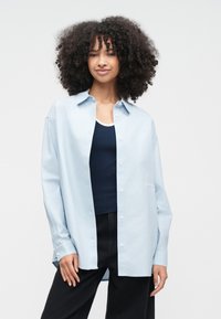 Ung person med lockigt hår som bär en ljusblå skjorta över en mörk linne och svarta byxor, stående mot en enfärgad bakgrund.