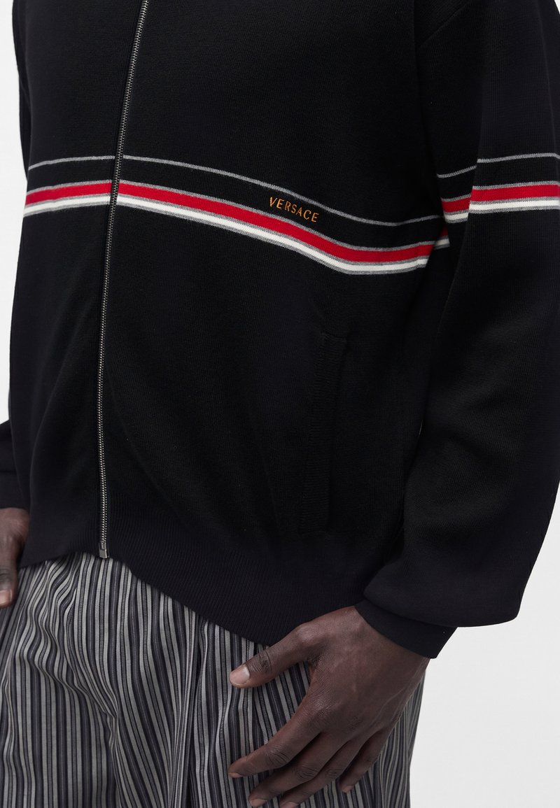 Pull noir zippé avec des rayures rouges, blanches et grises sur la poitrine et les manches, avec le texte « Versace », porté avec un pantalon gris rayé.
