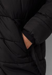 Schwarze Daunenjacke mit einem Reißverschluss-Taschen, gesteppter Optik und weichem Fleece-Bündchen, die eine glatte Textur und praktische Details betont.