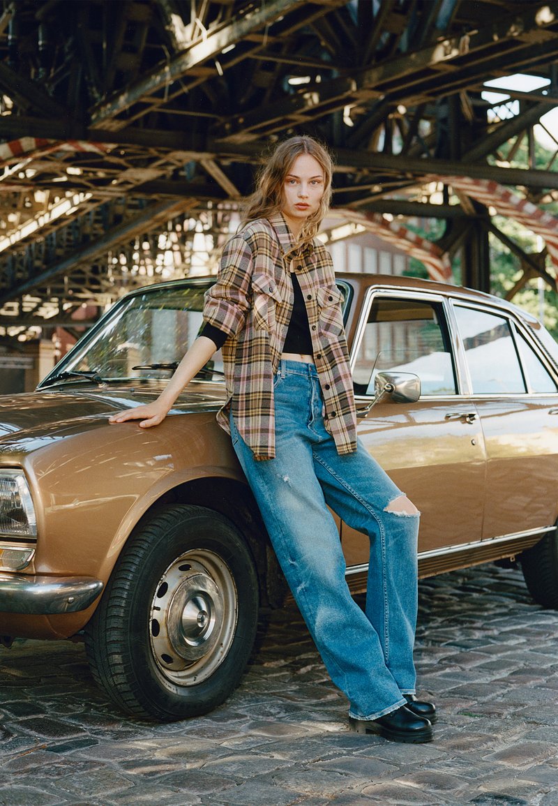 Camisa de flanela xadrez combinada com uma blusa preta cropped e jeans rasgados azul claro. Tamancos pretos, carro clássico com acabamentos em cromo ao fundo.