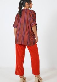 Blouse à coupe ample rouge avec des rayures violettes et des manches à motifs, assortie à un pantalon rouge vif. Les deux vêtements sont en tissu léger.