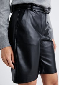 Mustat nahkashortsit, sileä pinta, laskokset edessä, vyölenkit ja sivutaskut. Yhdistetty harmaaseen neuleeseen, joka näkyy kalvosimissa.