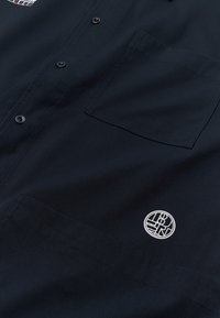 Chemise boutonnée sombre avec une poche poitrine et un petit logo rond blanc près du bas côté droit du tissu.