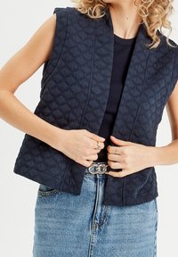 Frau in einer ärmellosen, marineblauen Steppweste über einem schwarzen Oberteil, mit einem blauen Jeansrock, einem silbernen Kettengürtel und Ringen an den Fingern.