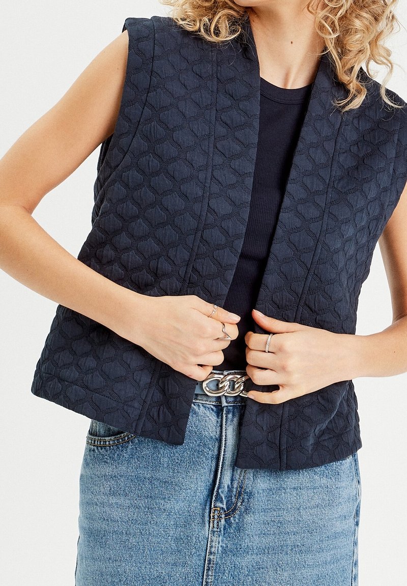 Femme portant un gilet matelassé sans manches bleu marine sur un haut noir, une jupe en denim bleu, une ceinture en chaîne argentée et des bagues aux doigts.