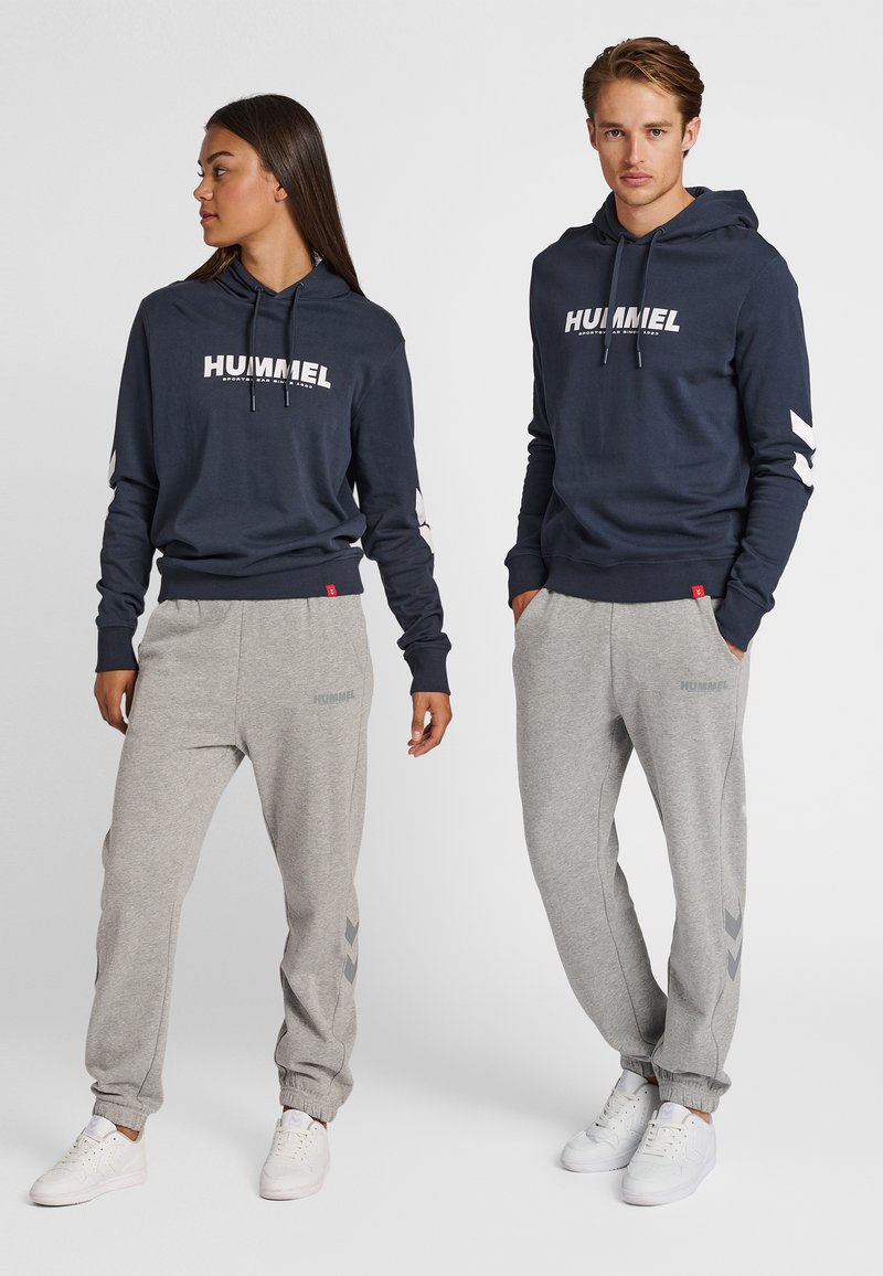 Mörkblå huvtröja med vit logotyp och dragsnören, kombinerad med grå mjukisbyxor med subtil märkning och chevron-detaljer vid mudden.