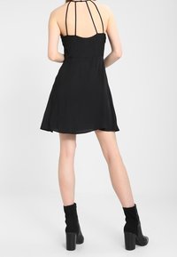 Black sleeveless dress with a flowy silhouette, featuring thin straps on the back, cut-out detail, and a smooth texture. Black ankle boots.