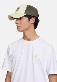 Gorra de color claro con una malla trasera verde, que presenta un parche gráfico de montaña. Se lleva con una camiseta blanca sencilla con un pequeño emblema amarillo.