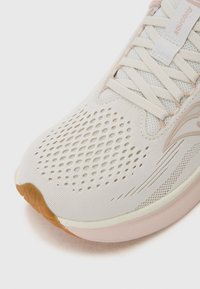 Chaussure de course légère avec une tige en mesh blanc cassé respirant, accent rose, semelle texturée et design perforé pour la ventilation.
