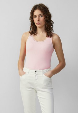 Roze geribbelde tanktop met een scalloped kant decolleté, gecombineerd met witte strakke jeans. Model staat met de handen in de zakken tegen een neutrale achtergrond.