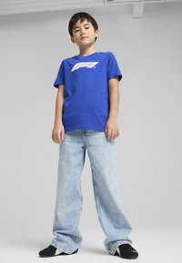 Modré bavlněné tričko s bílým logem F1, spárované se světle modrými širokými džíny. Černé boty dokreslují ležérní outfit.