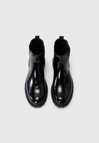Botas negras de charol hasta el tobillo con punta redondeada, paneles laterales elásticos y suela de goma texturizada. Equipadas con lengüetas traseras para facilitar el calzado.
