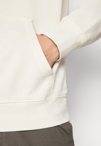 Felpa color crema con tasca frontale e polsini a coste. Il tessuto sembra strutturato, mostrando sottili dettagli di cucitura e una vestibilità rilassata.