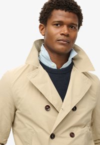 Beige dubbelzijdig trenchcoat met grote bruine knopen, gedragen over een marineblauwe gebreide trui en een lichtblauwe blouse met kraag.