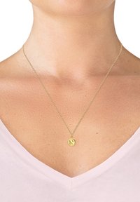 Guld vedhængs halskæde med et rundt charm gravert med et "N," hængende fra en delikat kæde. Baggrunden har en lysefarvet skjorte.