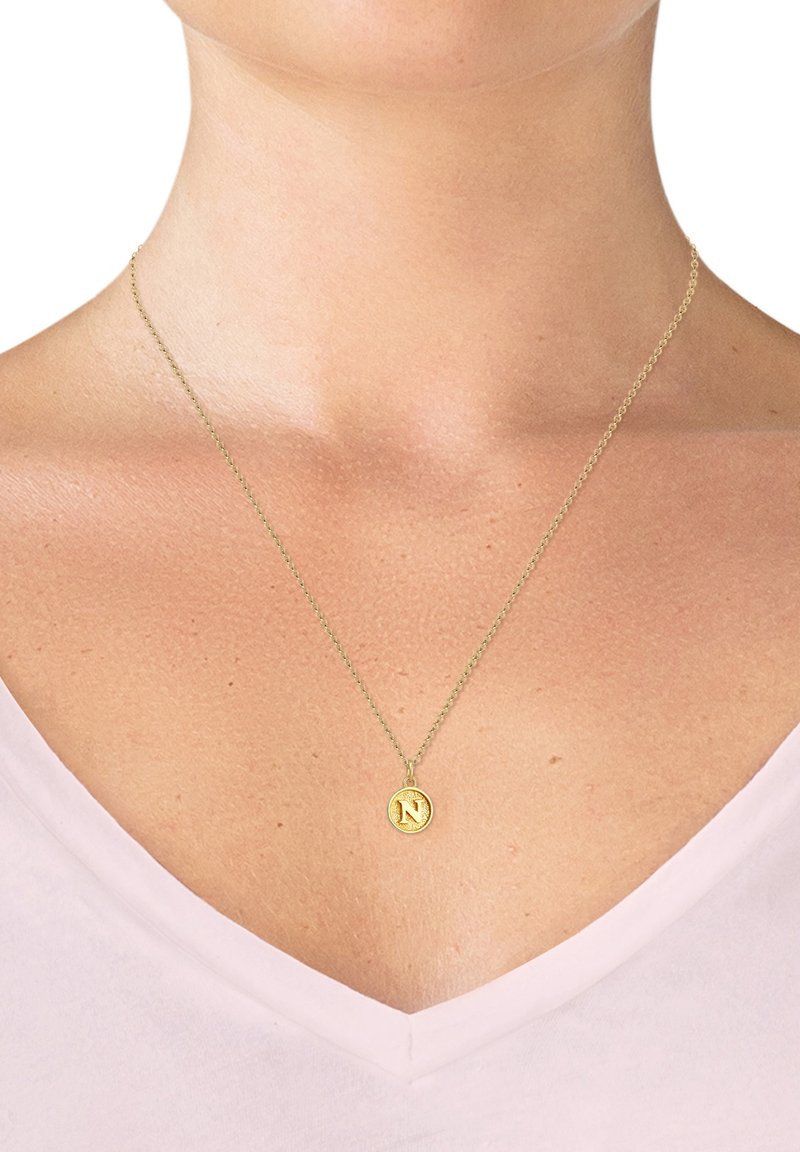 Guld vedhængs halskæde med et rundt charm gravert med et "N," hængende fra en delikat kæde. Baggrunden har en lysefarvet skjorte.
