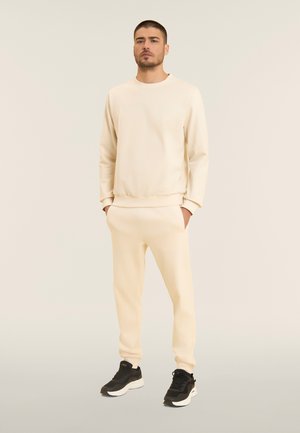 Mies seisoo kädet taskuissa, beige collegepaita ja sitä vastaavat verkkarit päällä, mustavalkoiset tennarit, beige tausta.