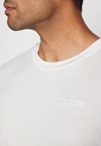 Weißes Rundhals-T-Shirt aus weichem Baumwollmaterial mit einem dezenten, geprägten "PeakPerformance"-Logo auf der Brust. Sanfte Textur, lockere Passform.
