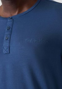 Mørkeblå bomulds henley-trøje med treknapslukning, med et subtilt præget logo og et sømformet X-detalje på kraven.