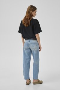 Sieviete ar sarkaniem matiem, nēsājot vaļīgu melnu T-kreklu, gaiši zilas džinsas ar aizmugurējām kabatām, melnu jostu un brūnas ērti uzvelkamas kurpes, stāvot un skatoties pa labi.