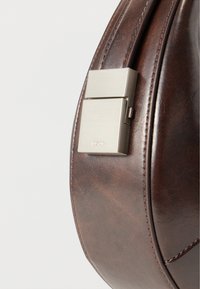 Κοντινό πλάνο ασημένιου ορθογώνιου κλεισίματος προσαρτημένου σε καμπύλη επιφάνεια από σκουρό καφέ δέρμα με εμφανείς ραφές.