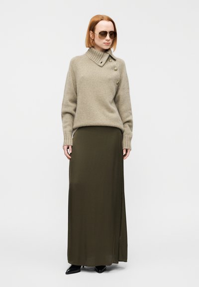 Μπεζ μάλλινο πουλόβερ με ψηλό γιακά και λεπτομέρεια κουμπιού, συνδυασμένο με μακριά φούστα σε ελιά πράσινο και μαύρα παπούτσια με μυτερή μύτη. Υποστηρικτικά γυαλιά ηλίου.