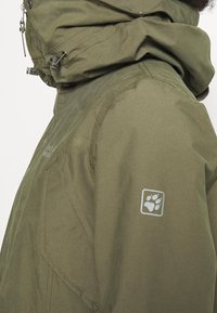 Olive green waterproof jacket with a high collar, adjustable hood, zip closure, and a paw print logo on the left sleeve. Textured fabric.