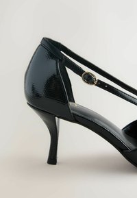Black patent leather high-heeled shoe with a pointed toe, ankle strap featuring a gold buckle, and a textured surface.