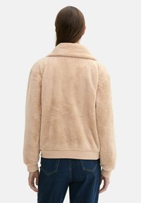 Chaqueta de forro polar beige con cuello con solapa, puños y dobladillo acanalados. El material es suave y texturizado, proporcionando una apariencia acogedora.