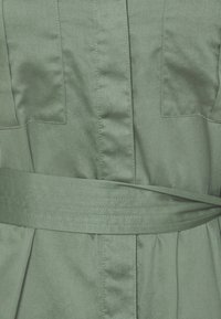 Abito in tessuto verde con una larga cintura cucita attorno alla vita e cuciture verticali visibili e tasche sul davanti.