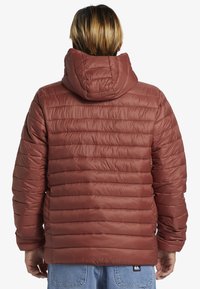 Rød isoleret puffersjakke med vandret quiltet mønster, påsat hætte og elastiske manchetter. Har logo på kanten.