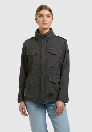 Fiatal nő sötétszürke, négy zsebbel ellátott munkakabátban és világoskék farmernadrágban, egyszínű háttér előtt állva.