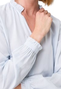 Helle blau gestreifte Bluse mit Rüschensaum am Halsausschnitt und an den Ärmeln. Aus weichem Stoff mit lockerer Passform und gerafften Details an den Ärmeln.