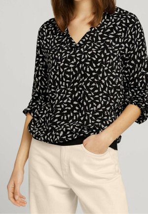 Femme portant un chemisier noir à manches longues avec des motifs de feuilles blanches et un pantalon crème, une main dans la poche et l'autre détendue le long du corps.
