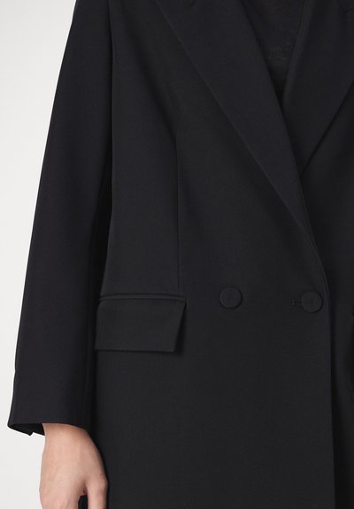 Blazer nero sartoriale doppiopetto con due bottoni e tasca con patta sul lato sinistro, indossato da una persona con mano parzialmente visibile.
