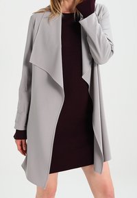 Jasnoszary płaszcz otwarty z drapowanym kołnierzem, warstwowym designem, długimi rękawami i gładką teksturą, noszony na dopasowanej ciemnoczerwonej sukience.